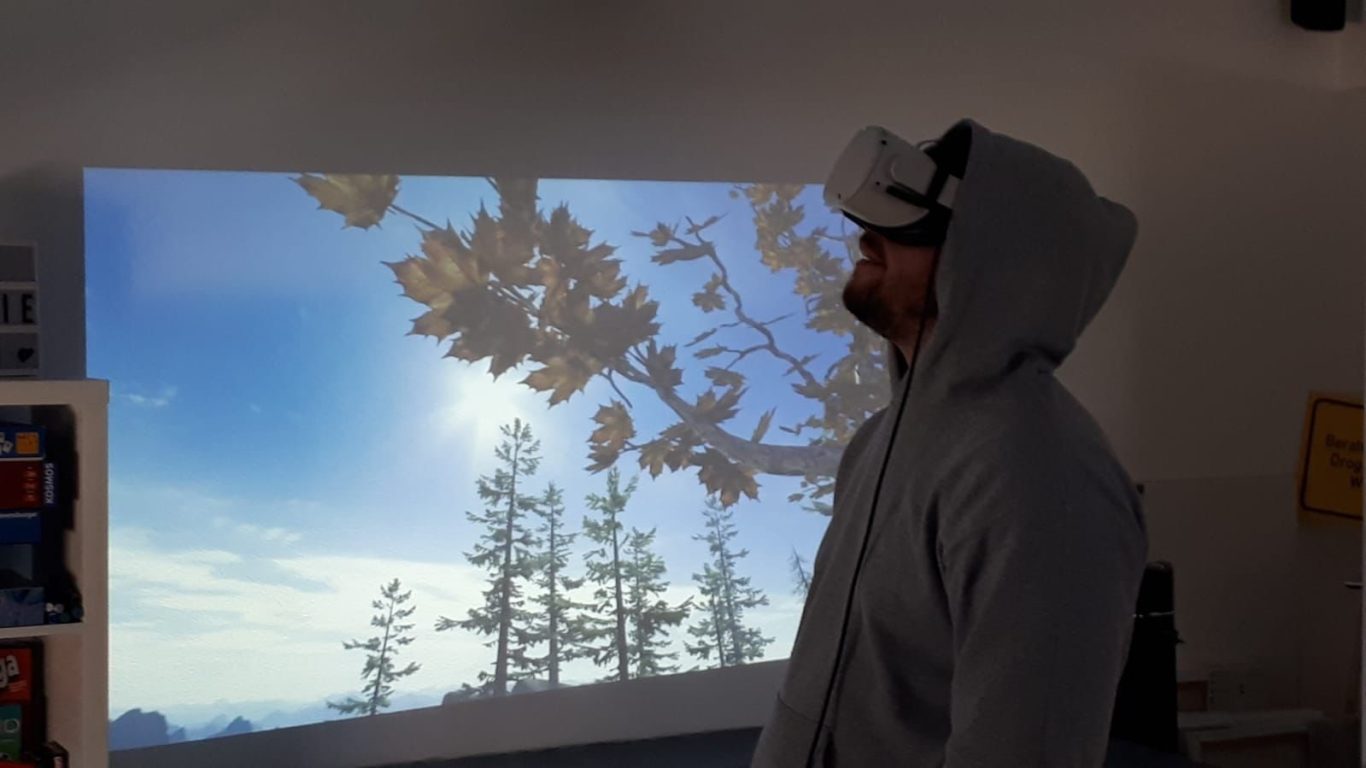 Mann in Hoodie steht vor Projektion einer Landschaft mit Bäumen und Himmel.