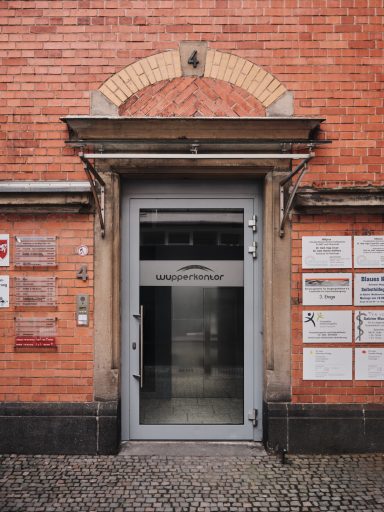 Graue Eingangstür in einem roten Ziegelgebäude mit Hinweistafeln daneben.